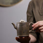 Grey-brown cylindrical teapot with bamboo decoration, held in two hands to show its size.