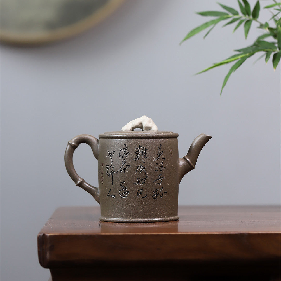 Grey-brown cylindrical teapot with bamboo decoration, opposite side showing calligraphy on a wooden table.