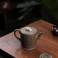 Grey-brown cylindrical teapot with bamboo decoration, opposite side viewed from above on a wooden table.