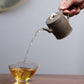 Grey-brown cylindrical teapot with bamboo decoration, pouring into a glass cup on a saucer.