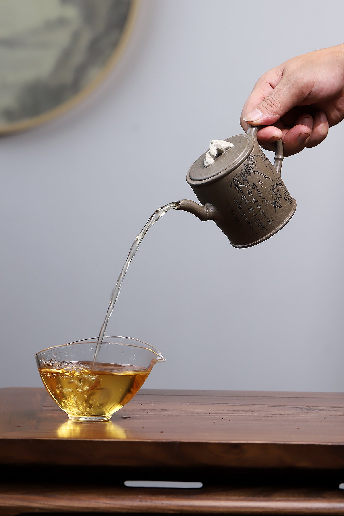 Grey-brown cylindrical teapot with bamboo decoration, pouring into a glass cup on a saucer.