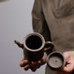 Grey-brown cylindrical teapot with bamboo decoration, open body and detached lid held in two hands.