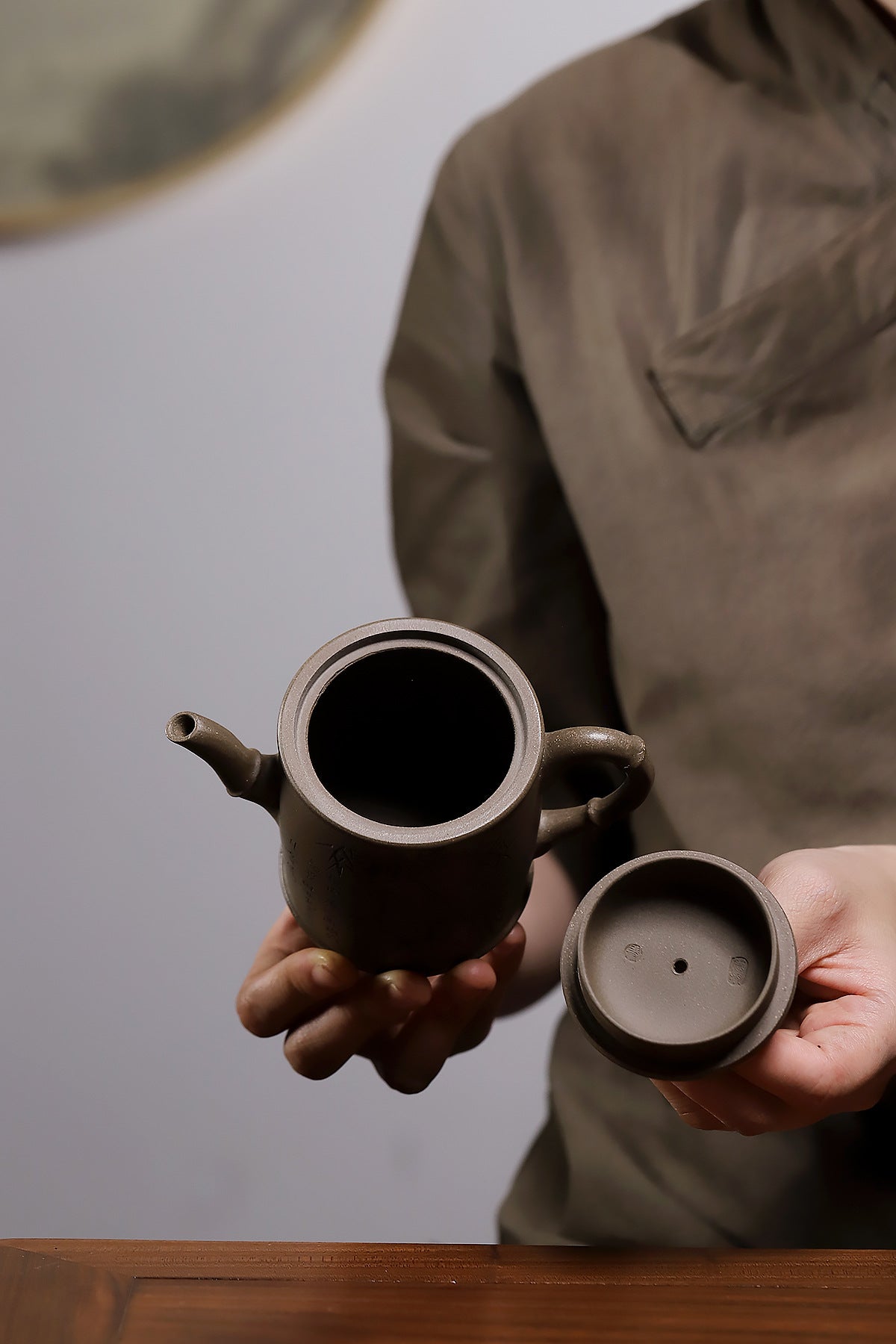 Grey-brown cylindrical teapot with bamboo decoration, open body and detached lid held in two hands.