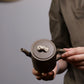 Grey-brown cylindrical teapot with bamboo decoration, top view while held in two hands.
